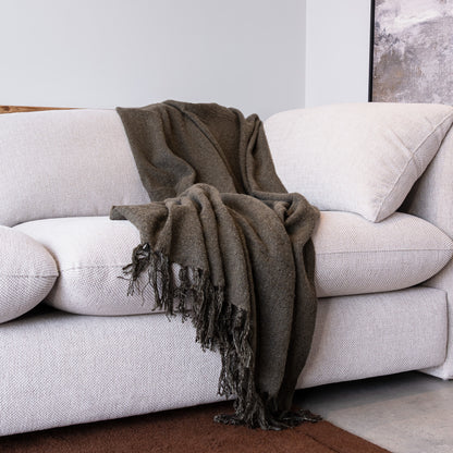 A dark brown textured throw blanket with fringed edges draped over a light beige fabric sofa in a modern living room setting.