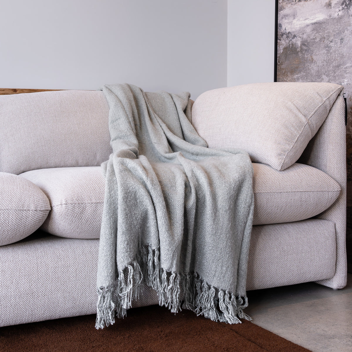 A light gray textured throw blanket with fringed edges draped over a cream-colored fabric sofa in a minimalist room setting.