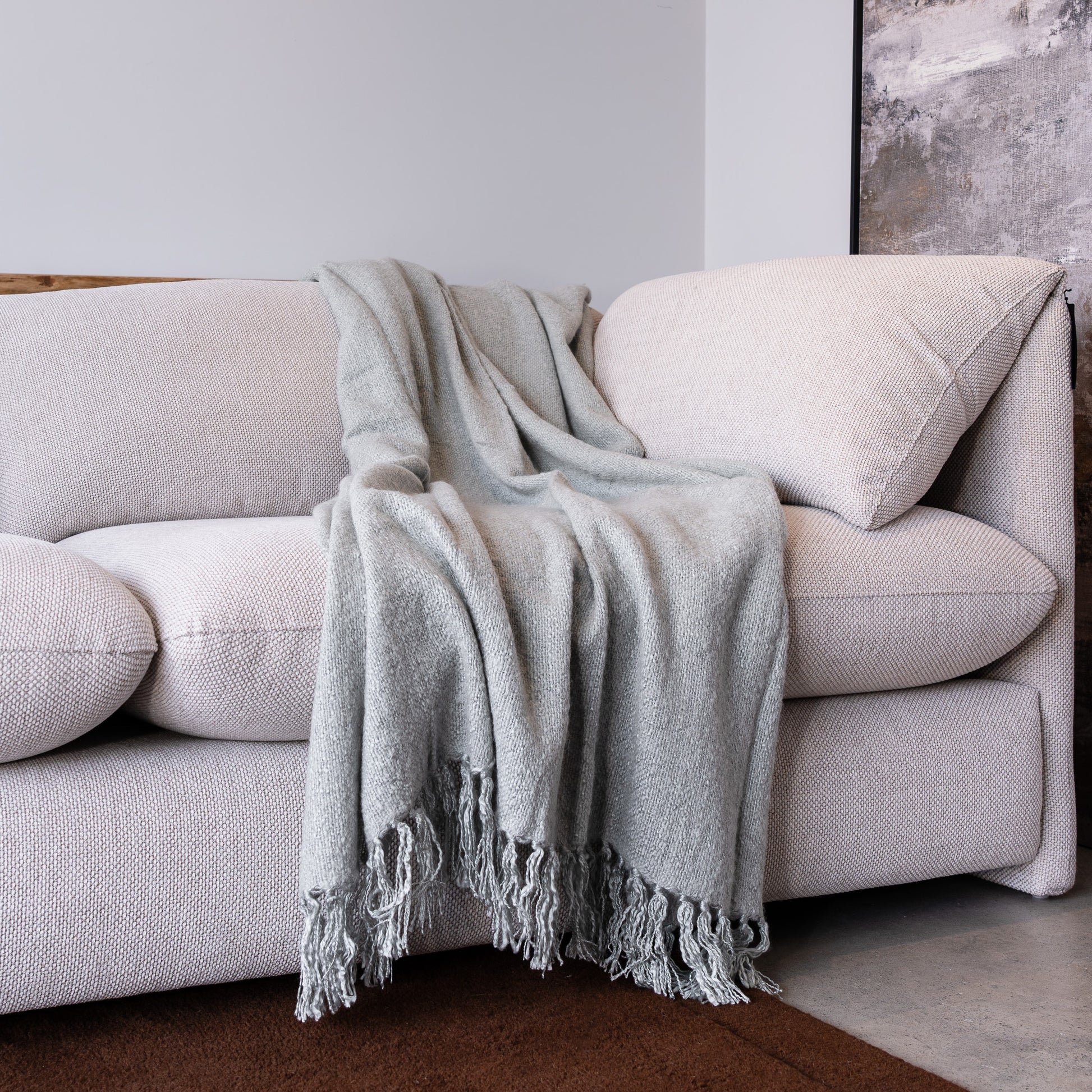 A light gray textured throw blanket with fringed edges draped over a cream-colored fabric sofa in a minimalist room setting.