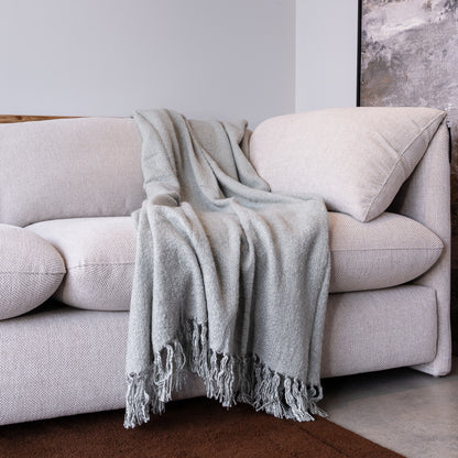 A light gray textured throw blanket with fringed edges draped over a cream-colored fabric sofa in a minimalist room setting.