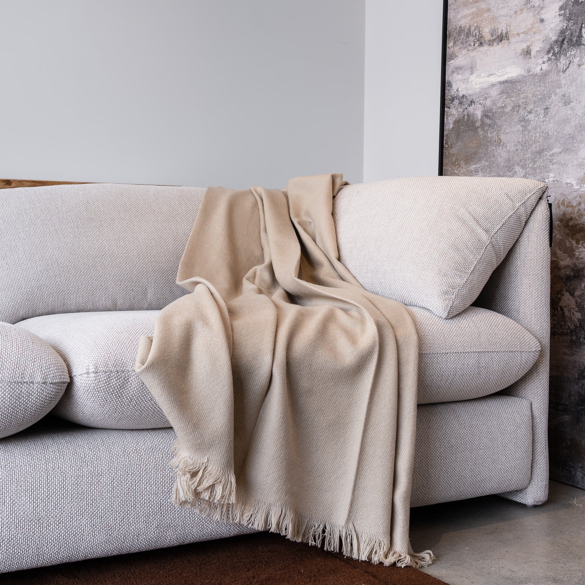 A light beige woven throw blanket with fringed edges draped over a light gray fabric sofa in a modern living room setting.