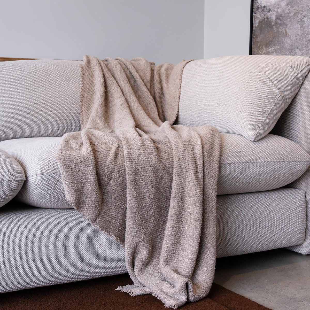 A textured beige throw blanket with fringed edges draped over a light gray fabric sofa in a minimalist living room setting.