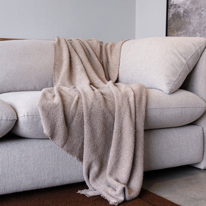 A textured beige throw blanket with fringed edges draped over a light gray fabric sofa in a minimalist living room setting.